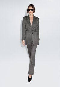 Szary dopasowany kostium z marynarką, która ma paski w talii, klapy typu notch oraz głęboki dekolt w kształcie litery V. Błyszczące czarne szpilki z podnosem w szpic i okulary przeciwsłoneczne.
