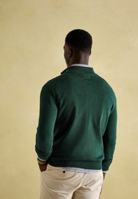 Zaļš adīts krekls ar ribotu apakšmalu un piedurknēm, uzvilkts virs pārbaudītas kreklam, ar augstu apkakles dizainu.