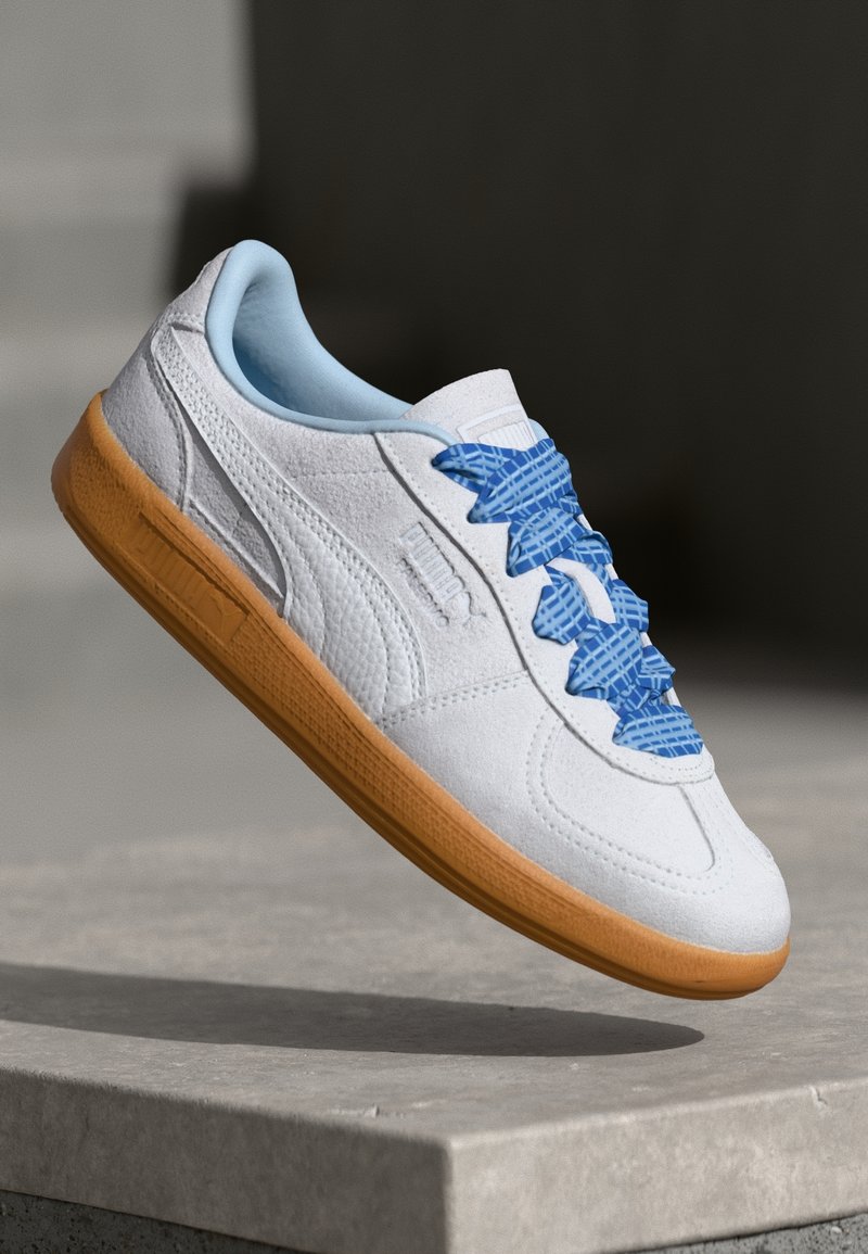 Baskets blanches Puma avec lacets à motifs bleus et semelle en gomme, montrées flottant au-dessus d'une surface en béton avec un fond sombre.