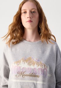 Sivý sveter s grafikou hory a textom. Mäkko tkanina s okrúhlym výstrihom. Dizajn obsahuje "COLORADO Mountains" v rôznych farbách.