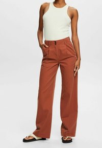 Pantalon large couleur rouille avec plis à l'avant, poches latérales et fermeture par bouton, associé à un débardeur blanc ajusté et des sandales.