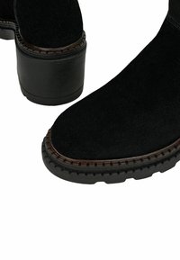 Czarne zamszowe botki za kostkę z zaokrąglonym noskiem, grubą gumową podeszwą i kontrastującym brązowym przeszyciem na krawędzi. Chropowata górna powierzchnia.