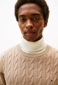 Maglione beige a maglia cable con un alto collo bianco. Pattern testurizzato e scollatura tonda. Piccola etichetta sul fondo a sinistra.