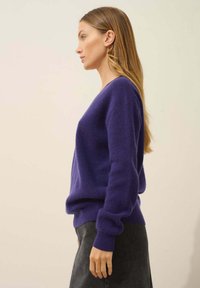 Lila stickad tröja med avslappnad passform, v-ringning, ribbade ärmslut och fåll, tillsammans med en mörkgrå kjol. Slät textur. Sido vy.