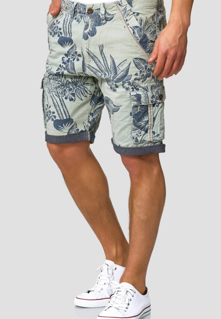 Shorts cargo vert clair avec un motif floral bleu marine, poches latérales, revers roulés et accents en métal. Baskets blanches assorties visibles.