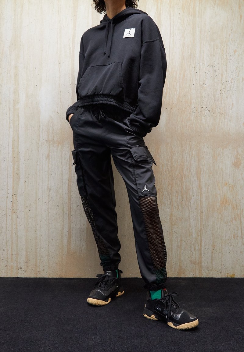 Zwarte hoodie met een voorzak en logo-patch, gecombineerd met zwarte cargo-broeken met netpanelen, aangevuld met zwarte sportschoenen.