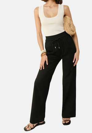 Femme portant un haut blanc sans manches, un pantalon noir à cordon, des sandales noires, des bracelets et tenant un sac tressé sous le bras.