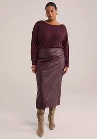 Burgundská halenka s dlouhým rukávem zkombinovaná s odpovídající sukní z umělé kůže se záhyby v diagonálním směru. Model nosí béžové vysoké boty po kolena.