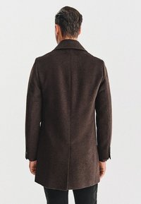 Brązowy wełniany płaszcz o dopasowanym kroju, z jednym rozcięciem z tyłu oraz klapami w wycięcie. Posiada mankiety na guziki i gładką teksturę.