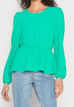 Femme portant un chemisier vert vif à manches longues avec une taille froncée et un jean bleu clair, debout les bras détendus.