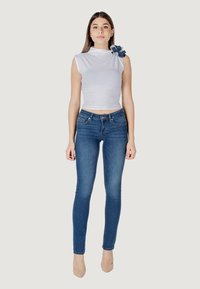 Top bianco aderente con un accento floreale sulla spalla sinistra, abbinato a jeans skinny blu e tacchi nudi. Texture liscia, senza motivi.