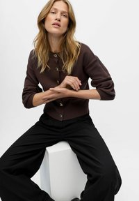 Rudas mezginys kardiganas su auksiniais mygtukais, turintis apvalią iškirptę ir 3/4 ilgio rankovėmis, derinamas su juodais plačiais keliais.