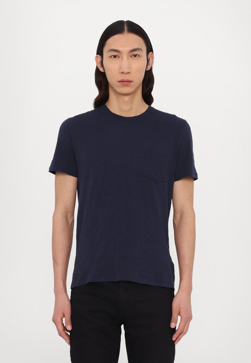 Camiseta de manga corta azul marino en una tela suave y texturizada, con cuello redondo y un bolsillo en el pecho, combinada con pantalones oscuros.