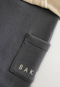 Ciemnoszara dzianina z małą prostokątną kieszenią z częściowo widocznym białym napisem "BAKE" i beżowym brzegiem u góry.