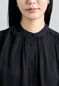 Blouse noire avec un col haut, un design plissé et des boutons à l'avant. Le tissu semble léger et a une finition douce et texturée.