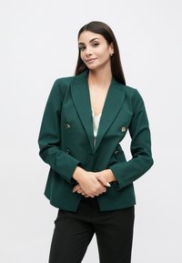 Blazer vert foncé croisé avec des boutons dorés, épaules structurées et coupe ajustée. Porté avec un haut de couleur claire et un pantalon noir.