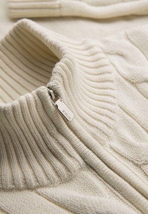Pull en maille de couleur crème avec un col montant côtelé et une fermeture éclair argentée partiellement tirée vers le bas.