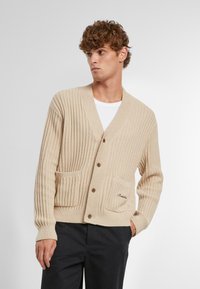Cardigan tricotat bej cu guler tip șal, două buzunare frontale, textură ribbed și nasturi din lemn. Broderie subtilă a logo-ului pe buzunar.