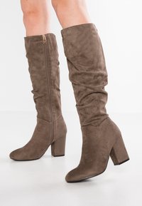 Brown suede knee-high boots with a slouchy design, featuring a side zipper and a chunky heel. Smooth texture with a rounded toe.