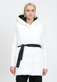 Weißer Kunstpelz-Weste mit Kapuze und schwarzem Gürtel, die eine weiche Textur und ein elegantes Design aufweist, getragen über einem weißen Langarmshirt.