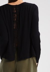 Schwarze Bluse aus strukturiertem Stoff, mit einem Spitzenbesatz entlang des Rückens, Knopfverzierungen und langen Ärmeln.
