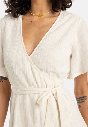 Femme portant une robe portefeuille blanche texturée avec manches courtes et taille nouée, collier en perles d'ambre, et tatouages visibles sur le bras.