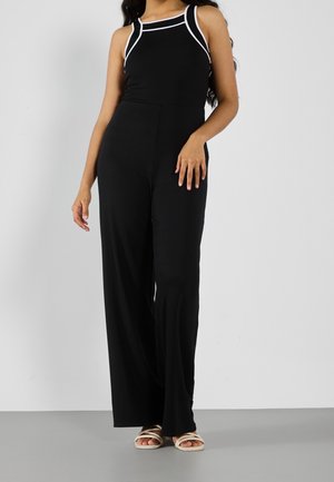 Femme portant une combinaison noire sans manches avec des finitions blanches et un pantalon à jambes larges, debout devant un fond gris uni.