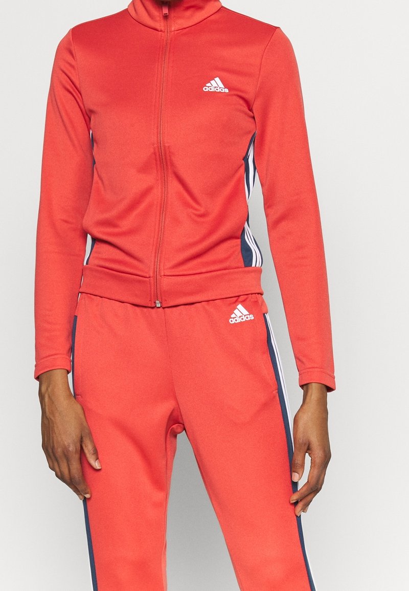 Personne portant un survêtement rouge Adidas avec une veste zippée intégrale et un pantalon assorti avec des bandes latérales blanches et bleu marine.