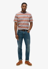 Stribet kortærmet skjorte i grå, orange og lilla, parret med blå jeans og brune snøresko. Har en rund halsudskæring og bælte.