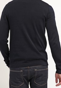 Osoba stojí zády k divákovi, oblečená v tmavě modrém svetru a tmavých džínách, čímž prezentuje neformální outfit.