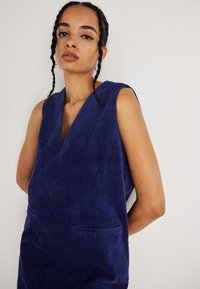 Bezramenné modré zamatové šaty s hlbokým výstrihom do V, dvoma prednými vreckami a štruktúrovanými ryhami, položené na neutrálnom pozadí.