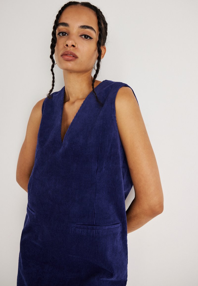 Bezramenné modré zamatové šaty s hlbokým výstrihom do V, dvoma prednými vreckami a štruktúrovanými ryhami, položené na neutrálnom pozadí.