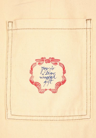 Pochette en toile naturelle avec une bordure rouge brodée et un texte manuscrit bleu : "La joie est un gros cadeau emballé." Design simple et texturé.