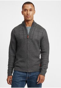 Grå strikket cardigan med glidelås fremme og sjalkrage. Har teksturert ribbing og et rutete mønster på den øvre fronten.