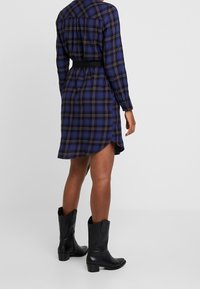 Rutigt skjortklänning i marinblått, lila och crème; har långa ärmar, krage och ett bälte i midjan; bärs med svarta ankelskor.