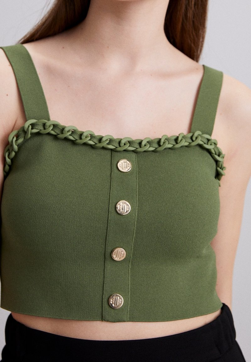 Débardeur court vert avec un détail de chaîne tissée le long de l'encolure. Comprend cinq boutons dorés sur le devant et un tissu texturé.