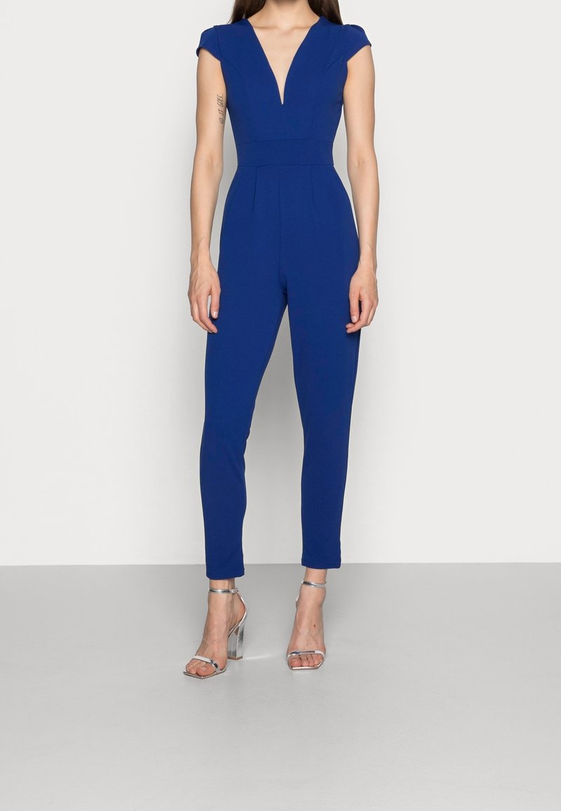 Kvinna i en figurnära, ärmlös blå jumpsuit med djup V-ringning, iförd silverfärgade högklackade sandaletter, som står mot en neutral bakgrund.