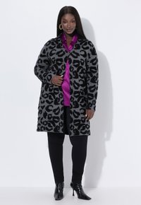 Grå leopardmönstrad kofta med svarta accenter, långa ärmar, öppen front, bärs över en glänsande lila topp och svarta leggings, ihop med svarta klackar.