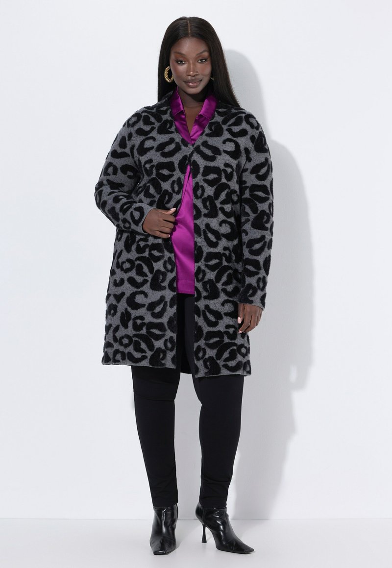 Grå leopardmönstrad kofta med svarta accenter, långa ärmar, öppen front, bärs över en glänsande lila topp och svarta leggings, ihop med svarta klackar.