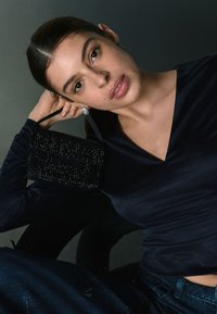 Svart pärlväska hålls i en hand, med en slank rem, kombinerad med en figursydd marinblå topp. Bakgrunden är neutral grå, vilket framhäver accessoarerna.