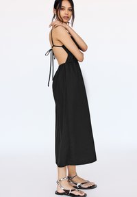 Robe sans manches noire avec un corsage ajusté, dos nu et taille froncée. Associée à des sandales noires et blanches à motifs.