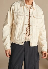 Chaqueta vaquera en color crema con dos bolsillos en el pecho, costuras visibles y dobladillo deshilachado. Lleva puesta sobre un top de punto texturizado y combinada con pantalones oscuros.