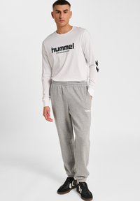 Langsleeve wit shirt met zwart "hummel" logo, gecombineerd met lichtgrijze joggingbroek met een elastische tailleband en zijzakken. Zwarte sneakers met accenten.