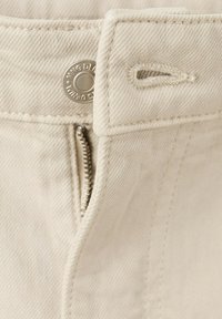Primo piano di tessuto denim beige che mostra un bottone di metallo, una zip e un occhiello sulla vita dei pantaloni.