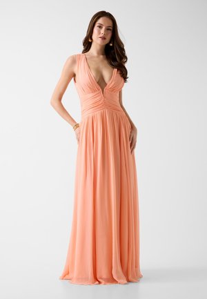 Kobieta w zwiewnej brzoskwiniowej sukni bez rękawów z głębokim dekoltem w serek, stoi z rękami w kieszeniach na jednolitym jasnym tle.