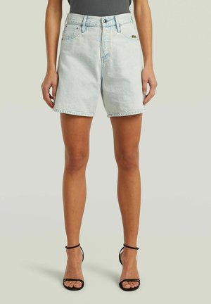 Vrouw draagt lichtblauwe high-waist denim shorts en zwarte sandalen met bandjes en hoge hakken, staand tegen een effen achtergrond.