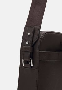 Tummanruskea teksturoitu nahkalaukku, jossa on säädettävä kangashihna, hopeinen metallisolki ja etutasku.