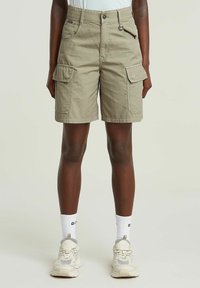 Shorts cargo en tissu beige clair. Comprend deux poches avant, deux poches cargo latérales et une fermeture par bouton. Accompagné de baskets et de chaussettes.