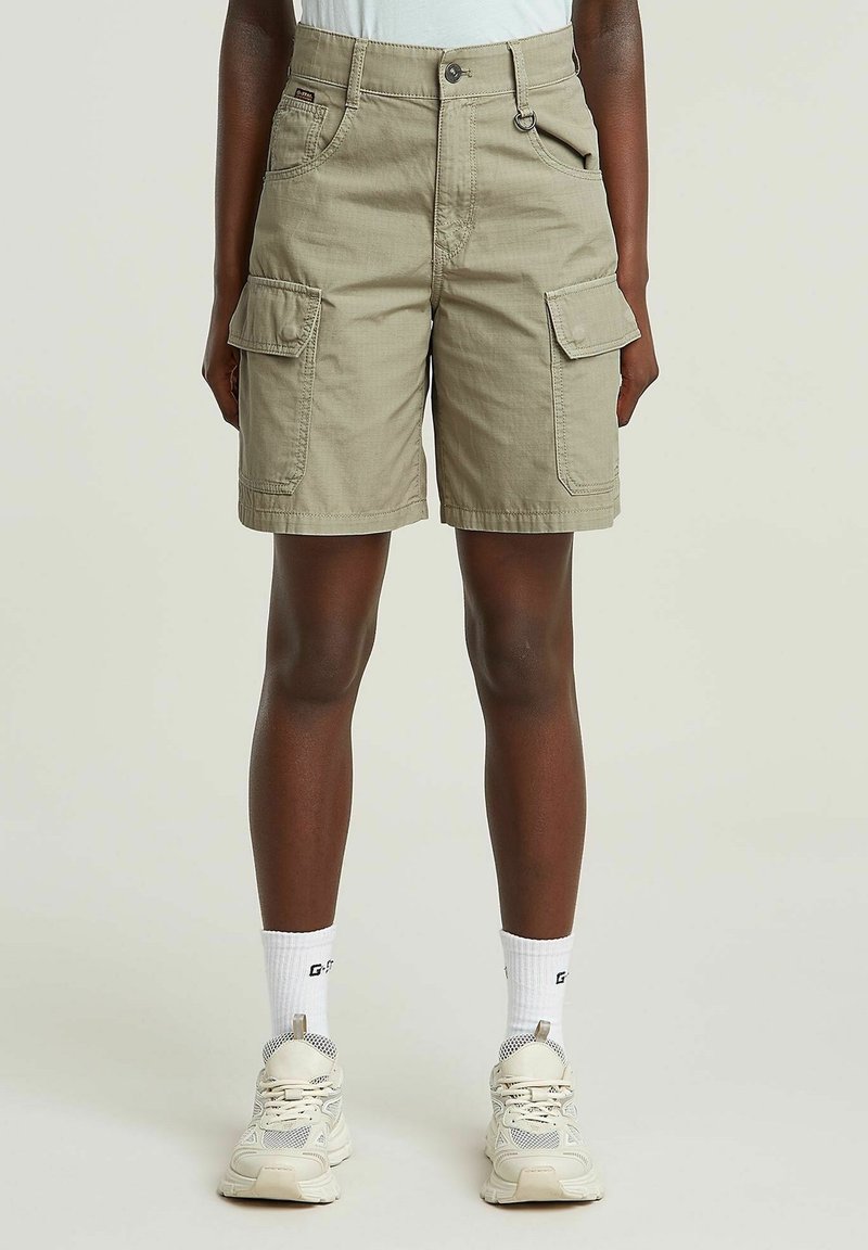 Shorts cargo en tissu beige clair. Comprend deux poches avant, deux poches cargo latérales et une fermeture par bouton. Accompagné de baskets et de chaussettes.