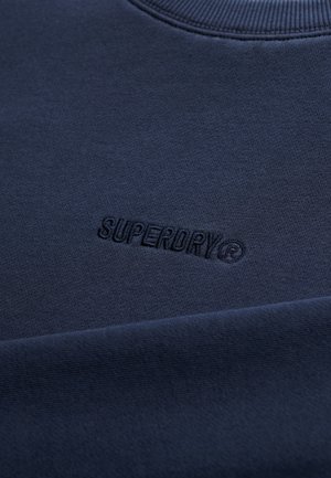 Granatowa bluza wykonana z miękkiej bawełny. Posiada prążkowany dekolt oraz tłoczony napis "SUPERDRY" na przodzie. Gładka tekstura.